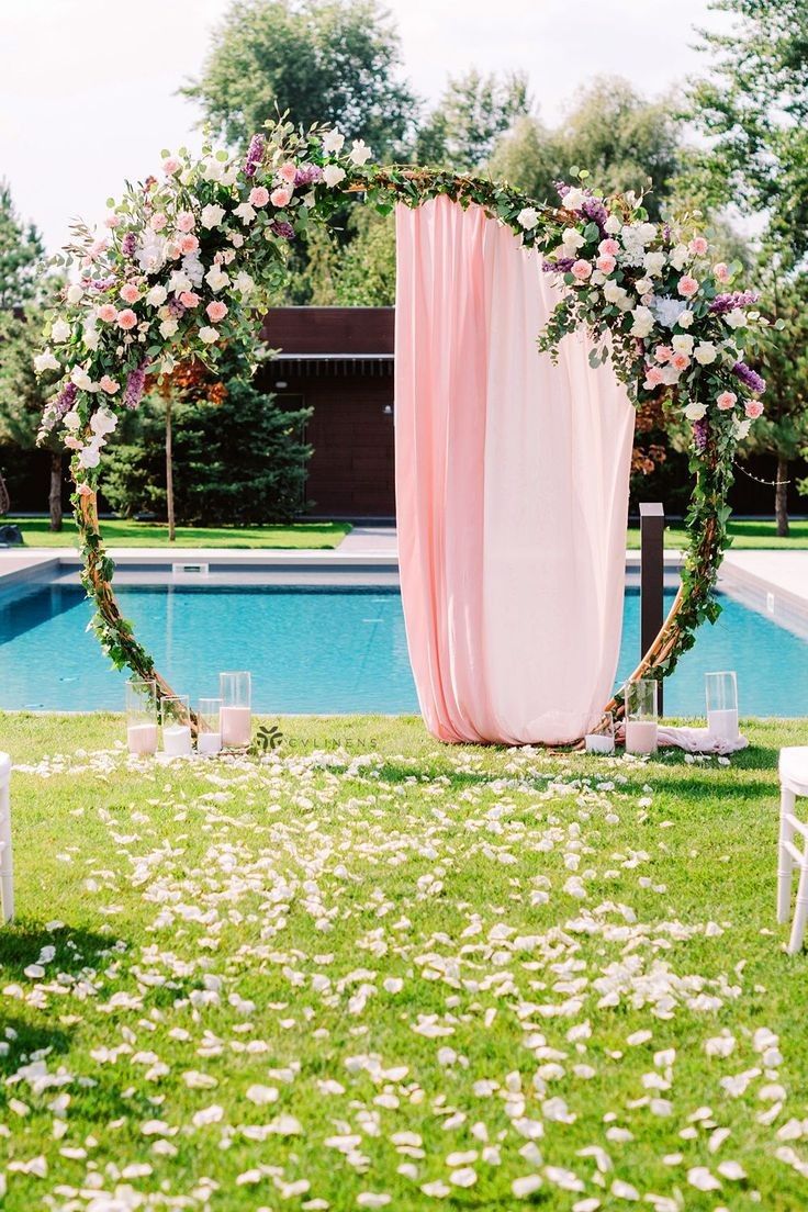 an outdoor ceremony setup with pink draping and white flowers on the grass next to a pool