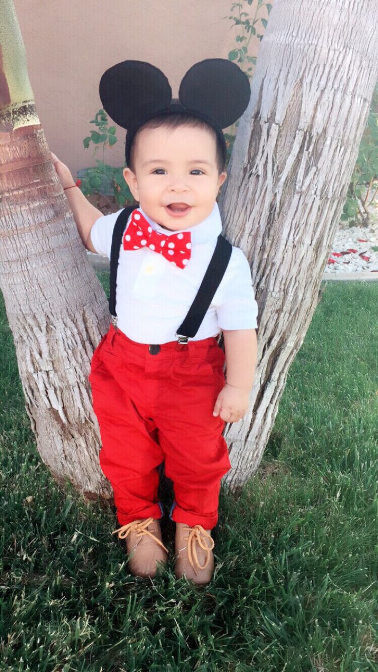 a little boy that is standing in the grass with a bow tie and suspenders