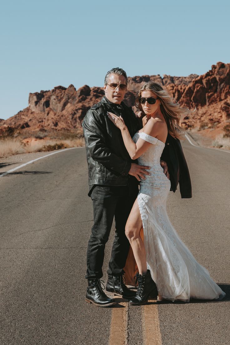 a man and woman standing on the side of an empty road in front of mountains