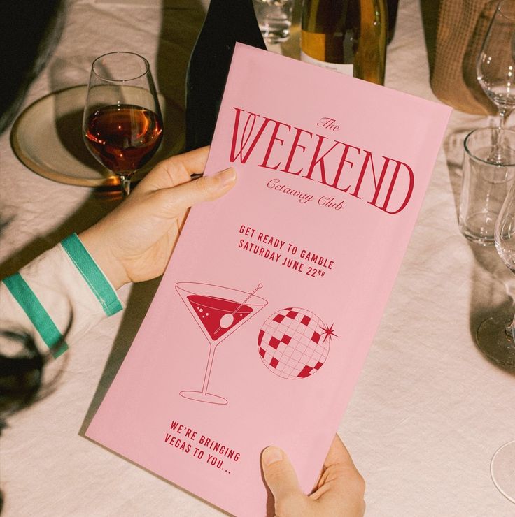 a person holding up a pink book at a table with wine glasses and plates on it