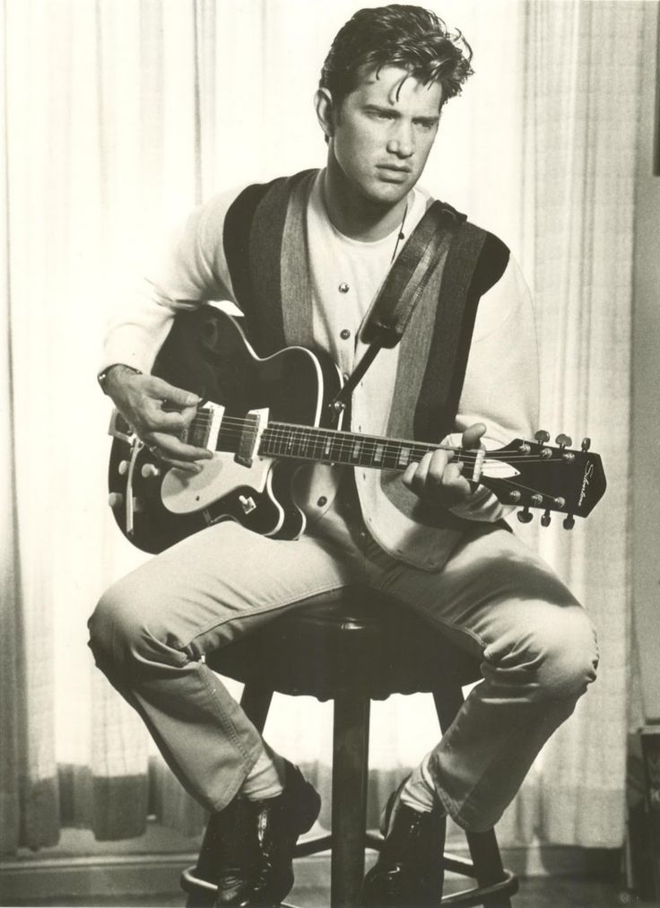 a black and white photo of a man sitting on a stool playing an acoustic guitar