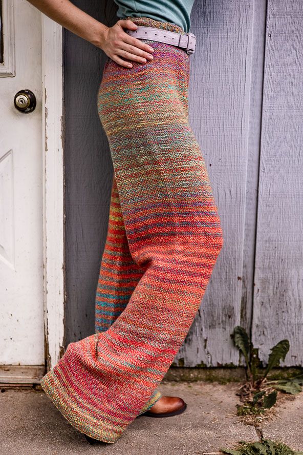 a woman leaning against a door with her hands on her hips wearing colorful pants and sandals