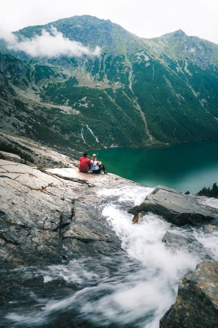 two people are sitting on the edge of a cliff overlooking a lake and mountain range