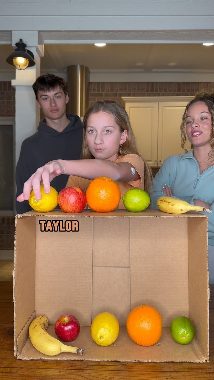 three people standing behind a cardboard box with fruit in it