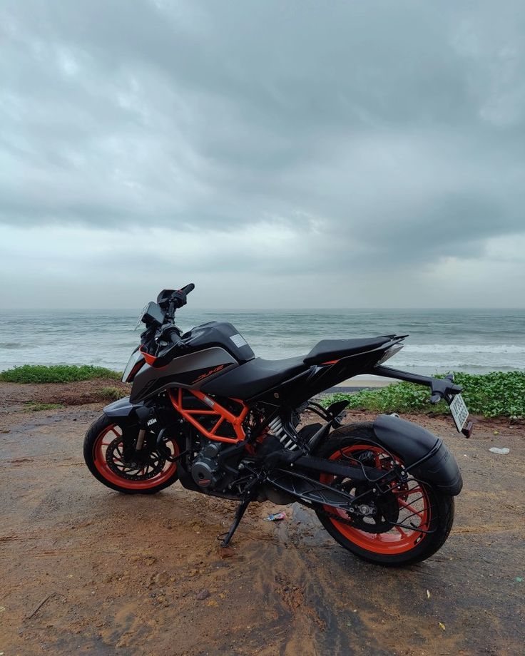 a black and orange motorcycle parked on top of a dirt field next to the ocean