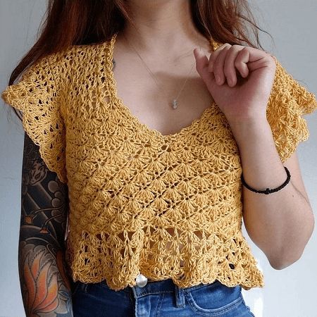 a woman wearing a yellow crochet top and blue jeans with tattoos on her arm