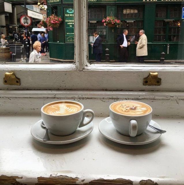 two cups of cappuccino sit on a window sill in front of people