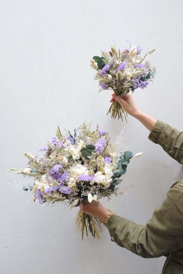 two hands holding flowers in front of a white wall with purple and white flowers on it