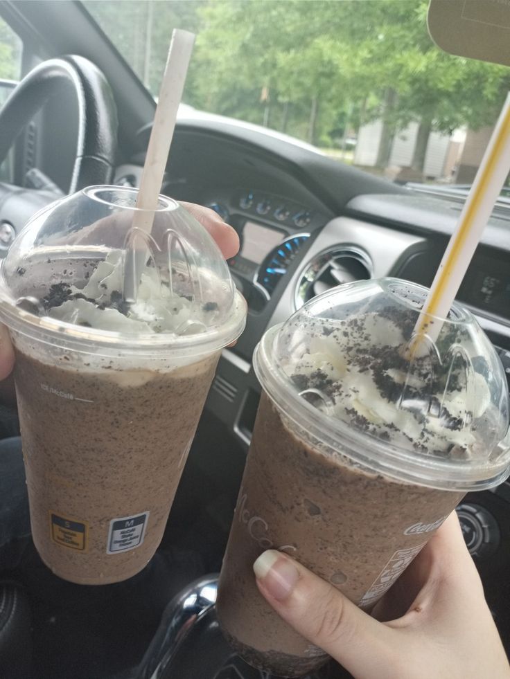 two people are holding up drinks in their hands while sitting in a car with the steering wheel behind them