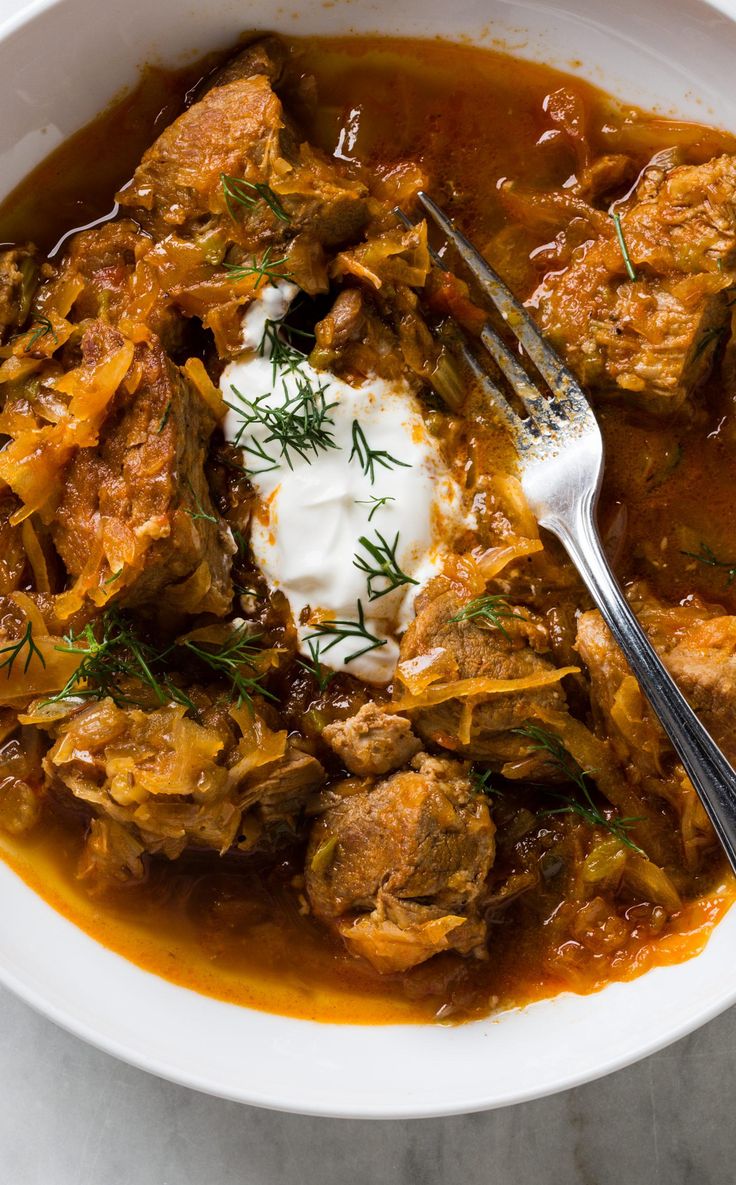 a white bowl filled with meat and vegetable stew next to a fork on top of a table