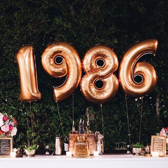 a table topped with lots of gold balloons and flowers next to a wall covered in greenery