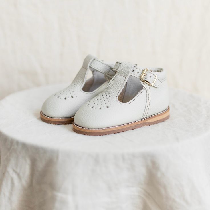 The Greta T-Strap in Fog is made of soft leather upper and lined in breathable leather which wicks away moisture. The structure of the shoe and the flexible rubber sole provide stability without weight to support your little one's early steps. We added an adorable teardrop perforation across the toe and a padded collar around the ankle. Perfect for everyday wear or special occasions. Bows by: The Little Design Co. Adjustable Leather Mary Janes For Spring, Classic T-strap Sandals With Round Toe For Spring, Classic Round Toe T-strap Sandals For Spring, White Closed Toe T-strap Sandals With Buckle, White Leather T-strap Sandals With Round Toe, Summer Closed Toe Mary Janes With Soft Sole, Spring Closed Toe Mary Janes With Stitched Sole, White Leather Mary Janes With Buckle Closure, White Closed Toe Mary Janes With Removable Insole