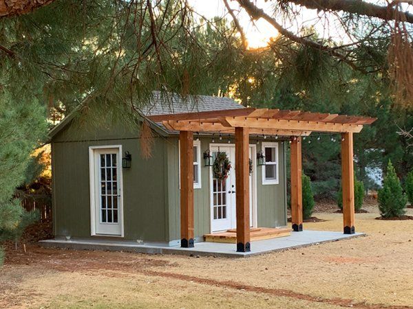 a small green house with a porch and covered in wood planks, surrounded by pine trees