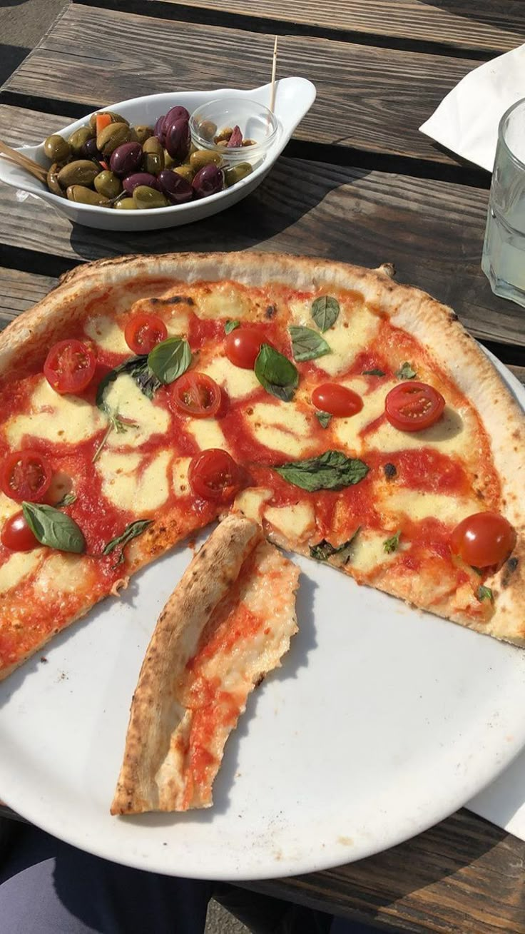 a pizza with several slices missing from it on a white plate next to bowls of olives