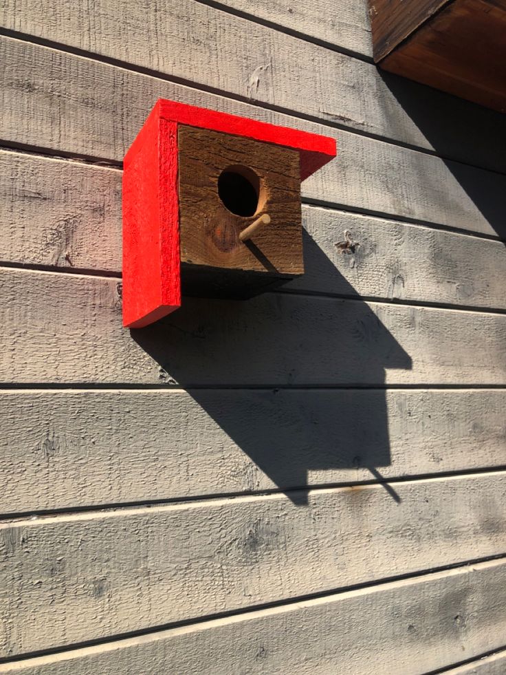 a red birdhouse hanging on the side of a gray wooden building with a window