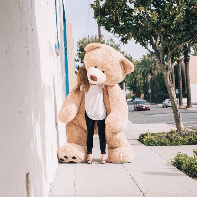 a woman standing next to a giant teddy bear on the side of a building with trees in the background