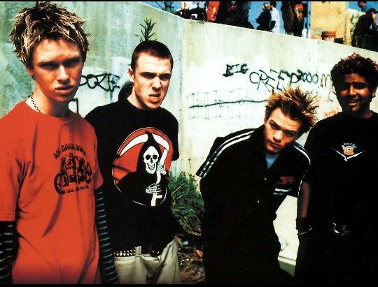 four young men standing next to each other in front of a wall with graffiti on it