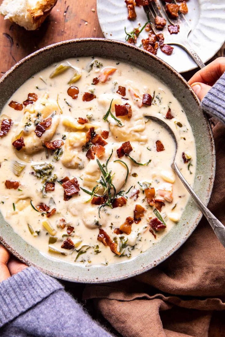 two hands holding a bowl of creamy potato soup with bacon and cheese on the side