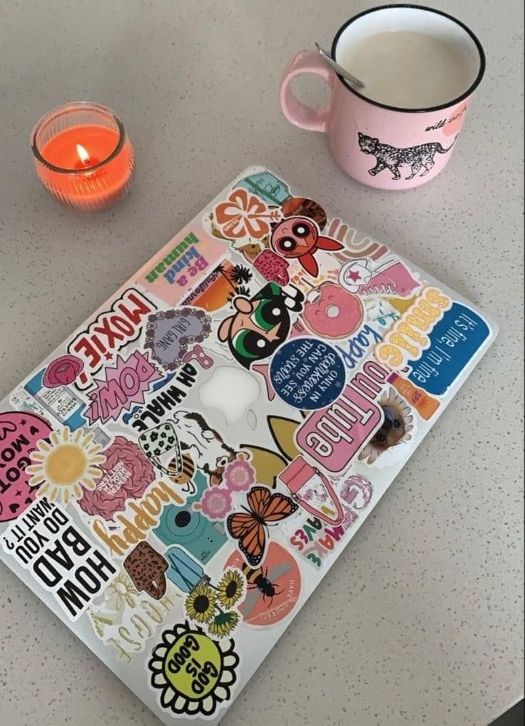 a cup of coffee next to a laptop with stickers on it and a lit candle