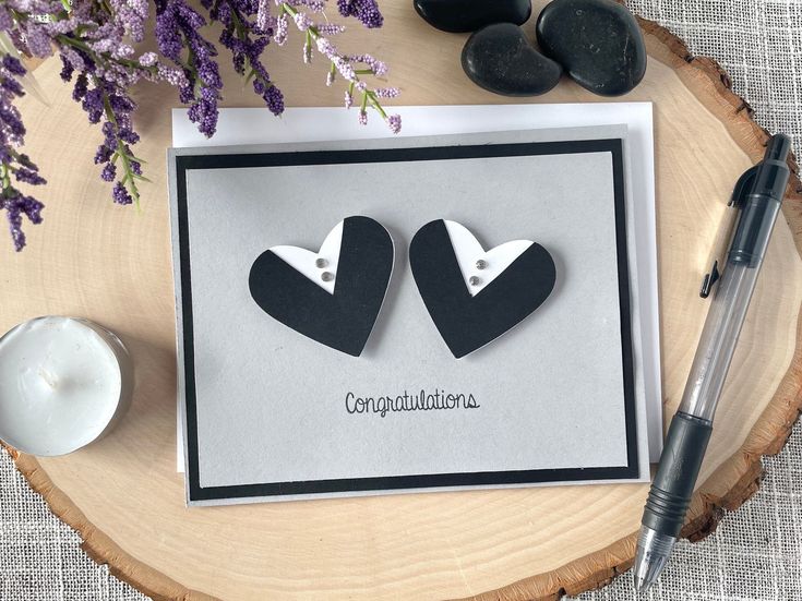 two black and white paper hearts on top of a card next to a candle, some rocks and flowers