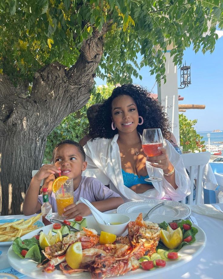 a woman and child sitting at a table with plates of food, drinks and fruit