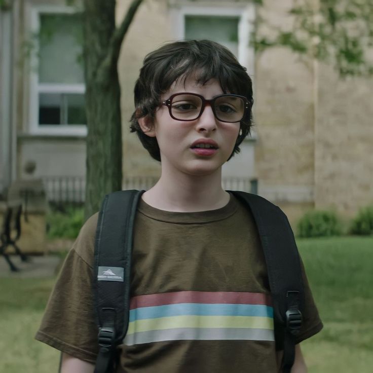 a boy with glasses and a backpack standing in front of a building, looking at the camera