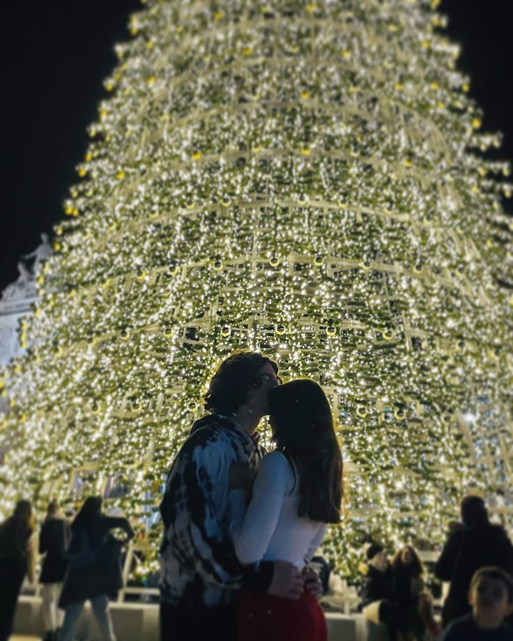 two people standing in front of a christmas tree at night with lights all around them