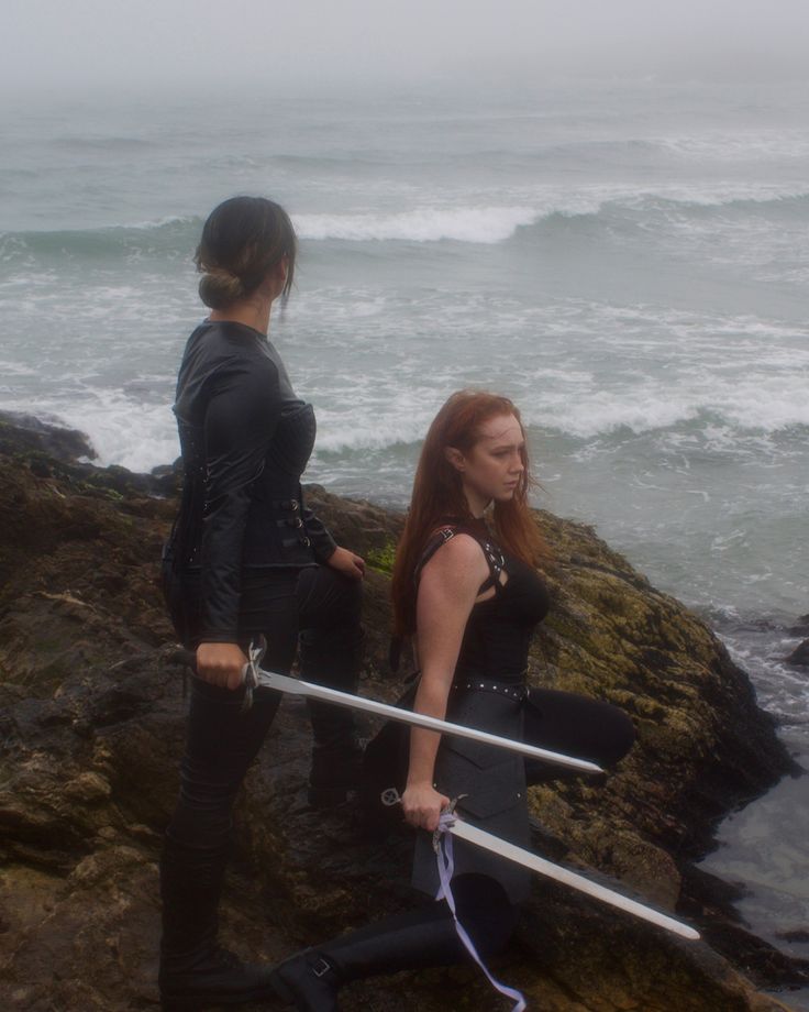 two women in wetsuits are standing on rocks near the ocean with their swords