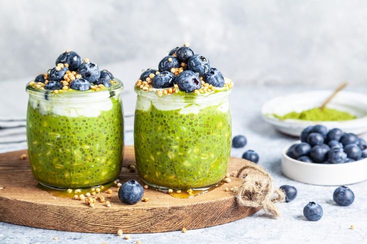 two jars filled with green smoothie and blueberries on top of a wooden board