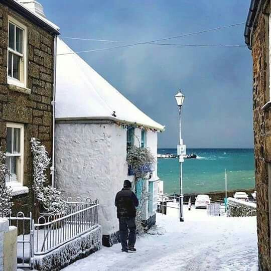 a man walking down a snow covered street next to the ocean with buildings on either side