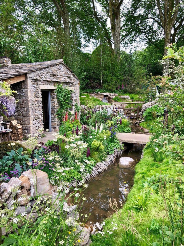 a small stone building sitting next to a stream in a lush green forest filled with flowers