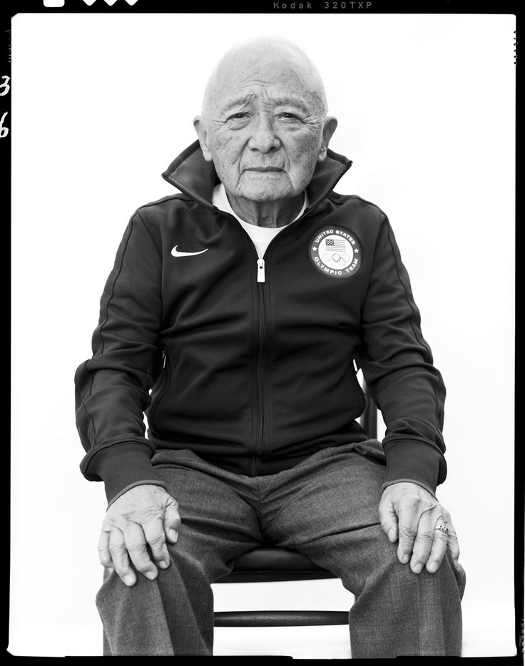 an old man sitting on top of a chair wearing a black jacket and grey pants