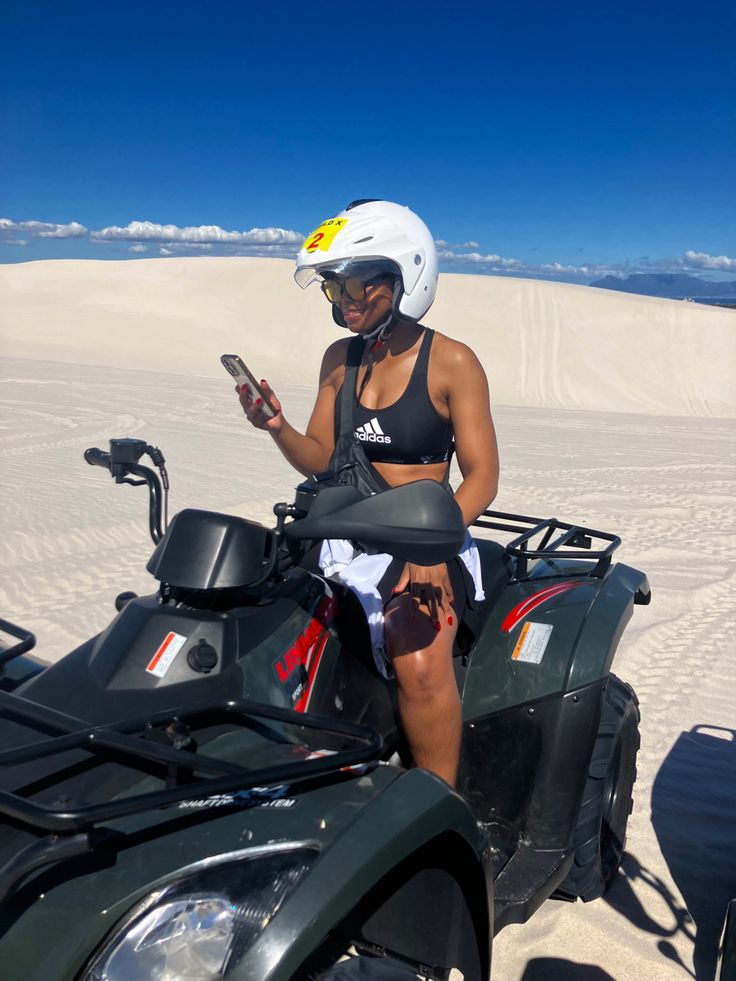 a woman sitting on top of an atv in the sand with her cell phone up to her ear