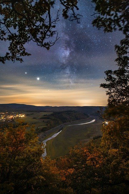 the night sky is lit up with stars and clouds above trees, overlooking a body of water