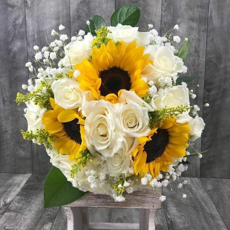 a bouquet of sunflowers and white roses sits on a wooden stand against a gray background