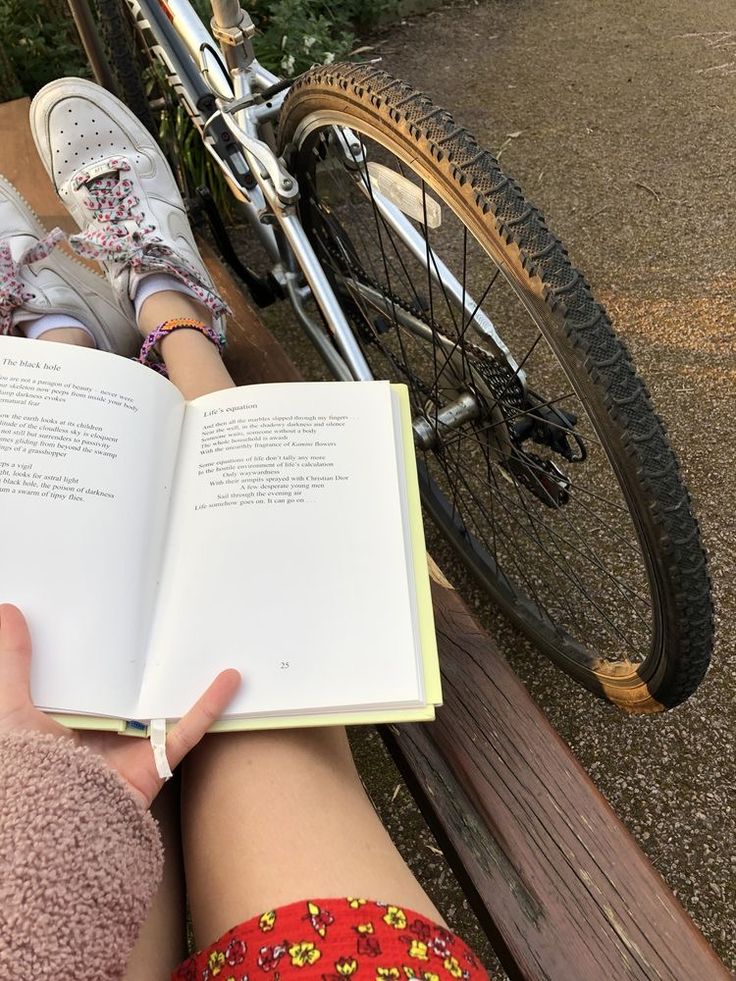 a person sitting on a bench reading a book next to a bicycle with their feet propped up