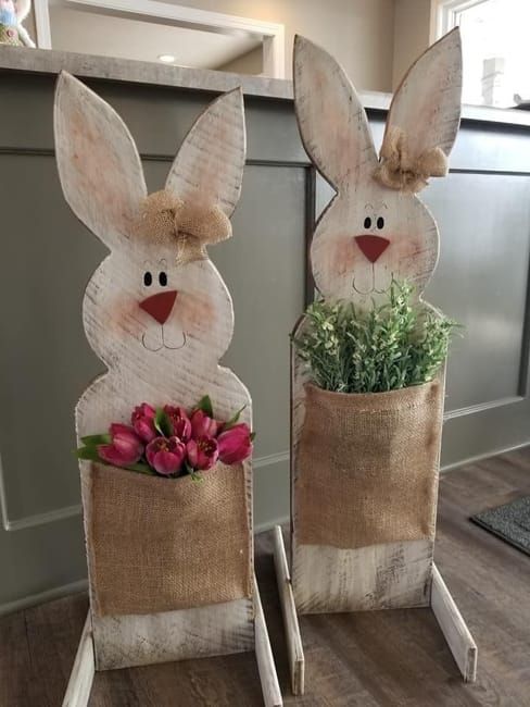 two wooden rabbits sitting on top of each other with flowers in their lap and one is holding a potted plant