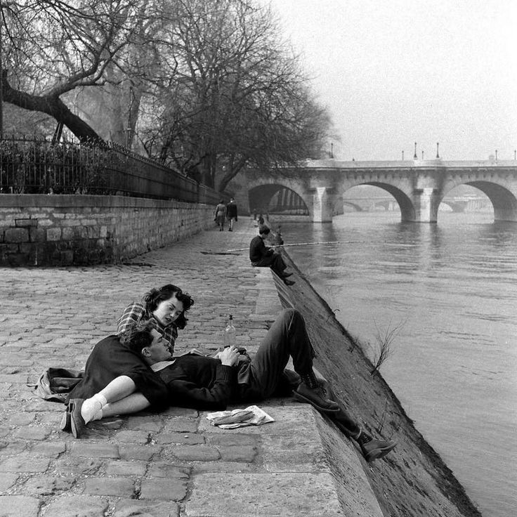 black and white photograph of two people laying on the ground next to water with bridge in background