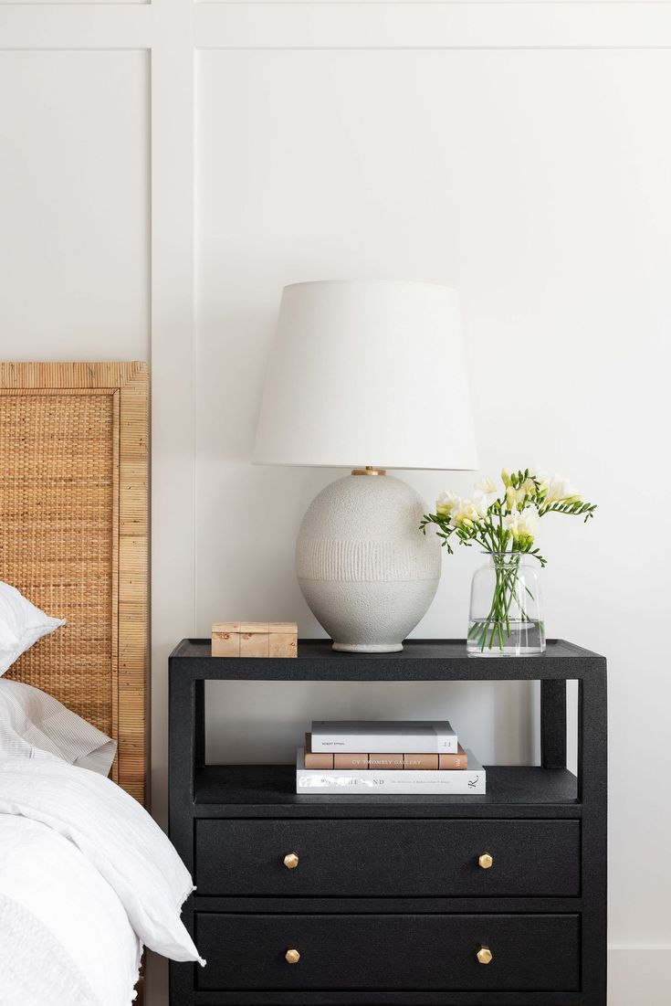 a nightstand with a lamp, books and flowers on it next to a white bed