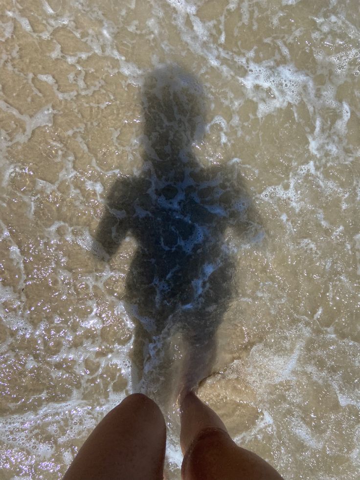 a person standing in the water with their shadow on the sand and behind them is a body of water