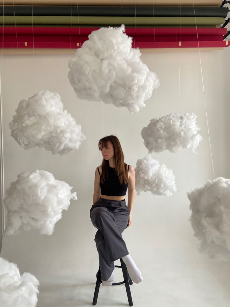 a woman is sitting on a stool in front of some clouds hanging from the ceiling