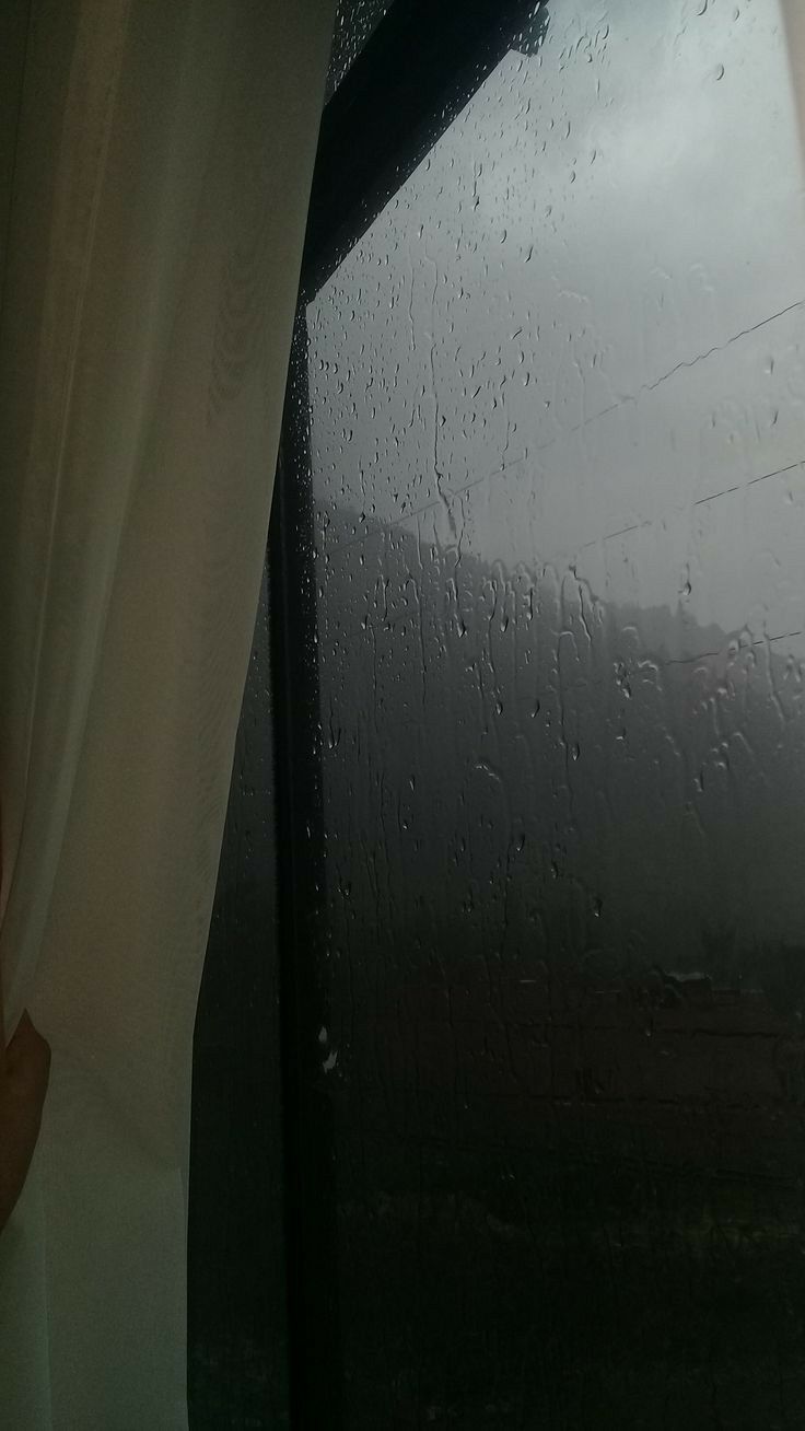 a hand holding an open curtain in front of a window with rain drops on it