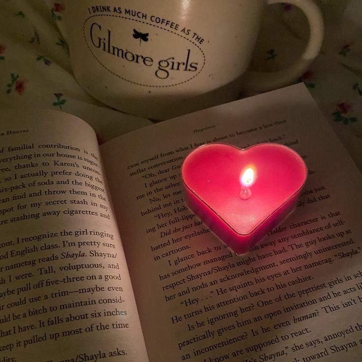 a pink heart shaped candle sitting on top of an open book next to a coffee mug