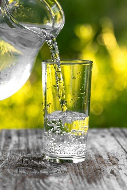 a pitcher pouring water into a glass on top of a wooden table with grass in the background