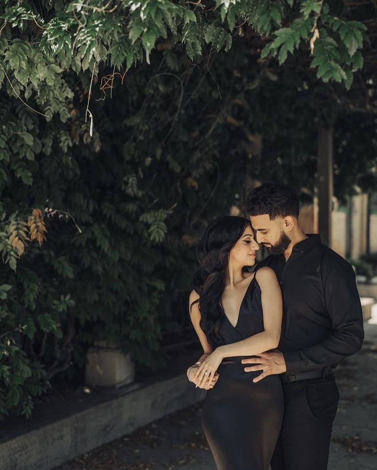 a man and woman standing next to each other in front of a tree with their arms around each other