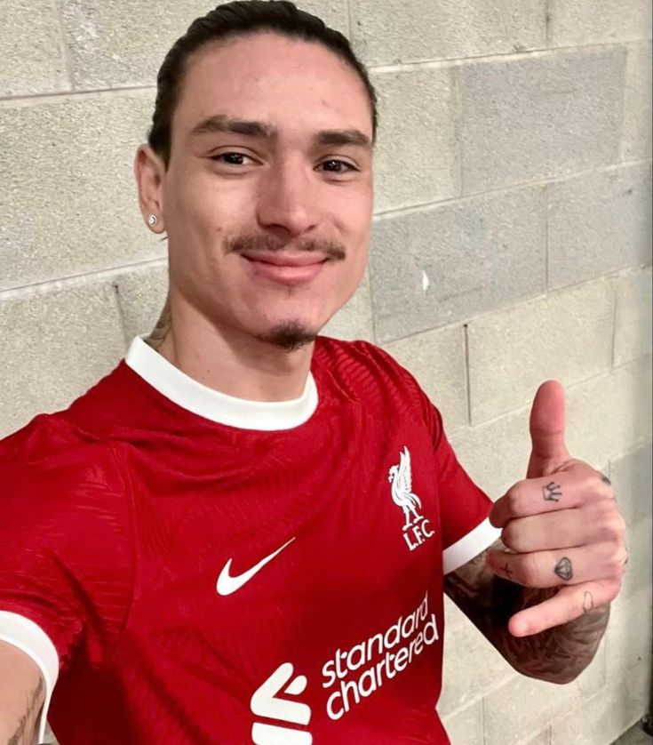 a man giving the thumbs up sign in front of a brick wall wearing a red liverpool shirt