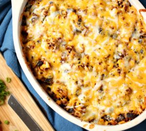 a casserole dish with cheese and other toppings on a blue cloth next to a wooden spatula