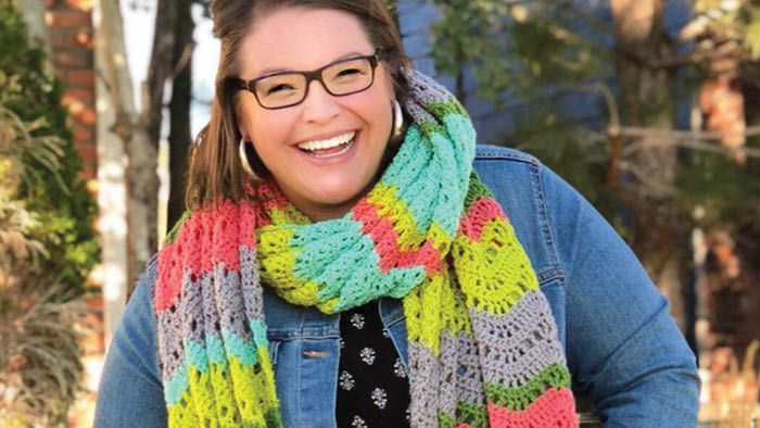 a smiling woman wearing glasses and a multicolored knitted scarf in front of trees