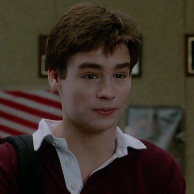 the young man is wearing a maroon sweater and white shirt with an american flag in the background