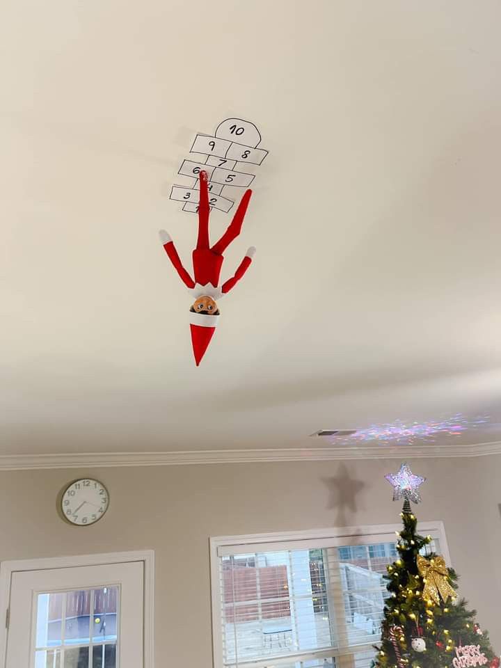 an elf hanging from the ceiling in front of a christmas tree and clock on the wall